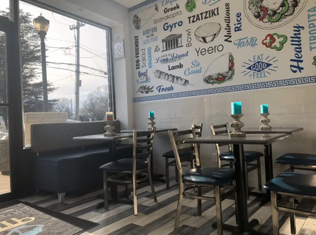 Inside Pita Bowl in Lindenhurst: blue booth seating and bistro tables under a hand-drawn mural of Greek foods and words like Yeero, Tzatziki and Made in Long Island