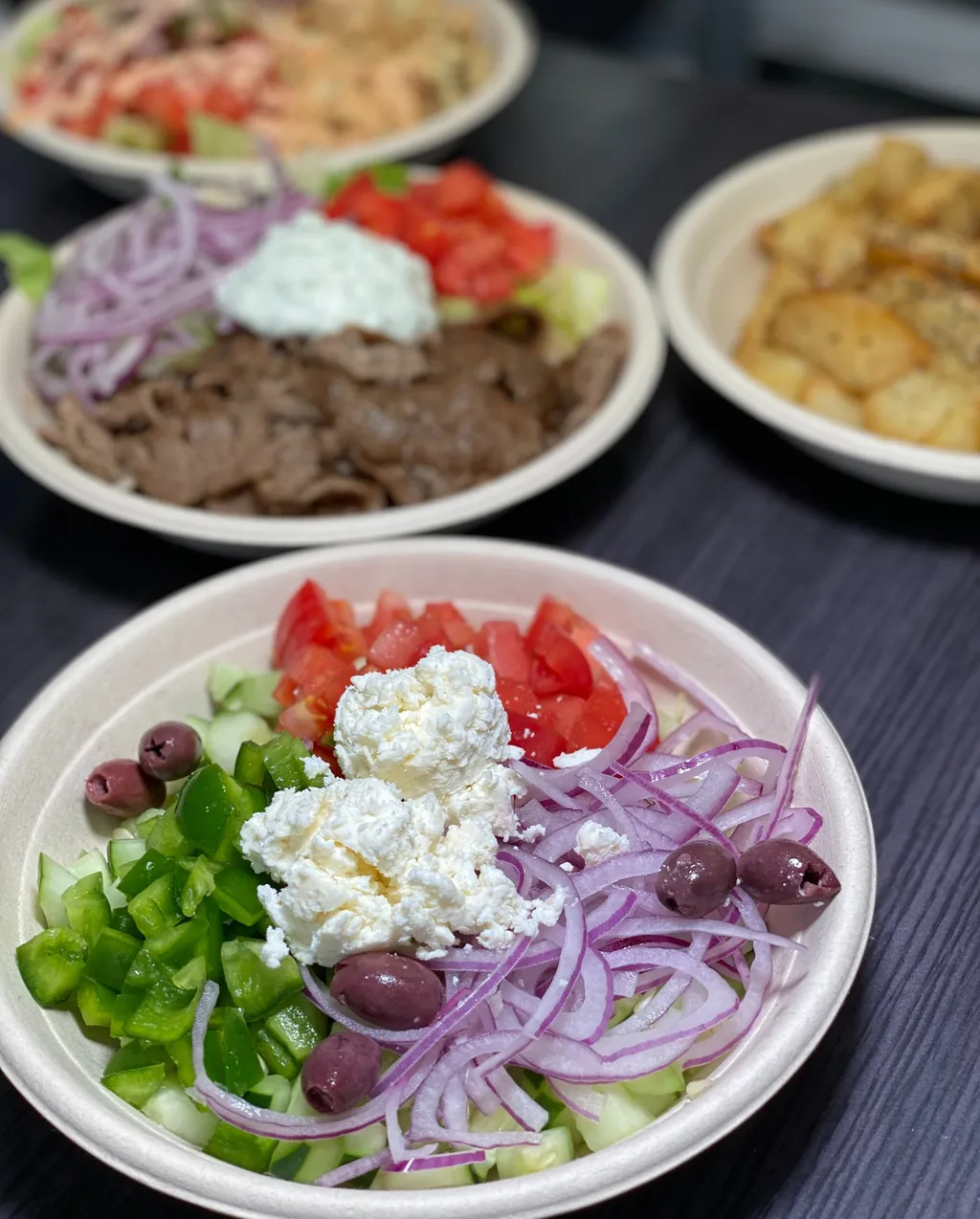 A gyro rice bowl with feta and lemon potatoes beside a Greek salad bowl with olives, green pepper and feta