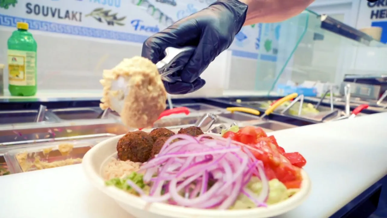 A Pita Bowl team member adds hummus to a falafel bowl with red onion and tomato at the build-your-own line, in front of the Greek mural